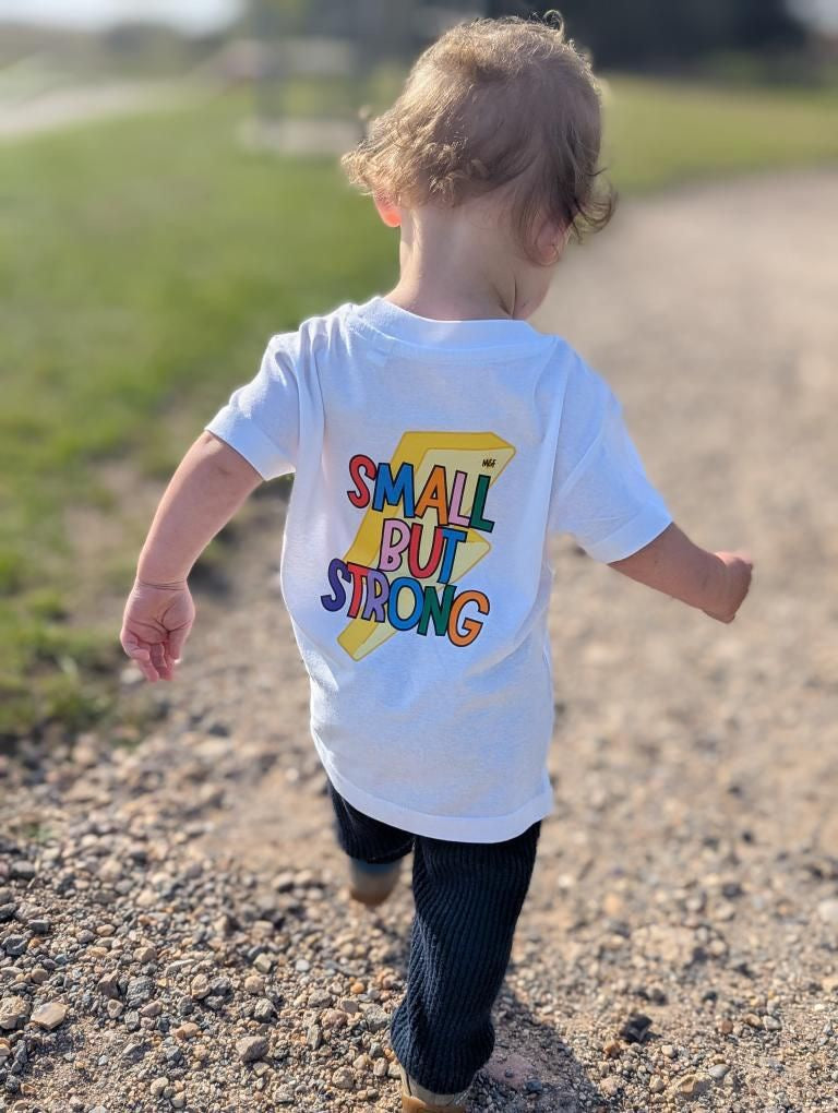 Child wearing a white t-shirt with 'Small But Strong' text, running outdoors.