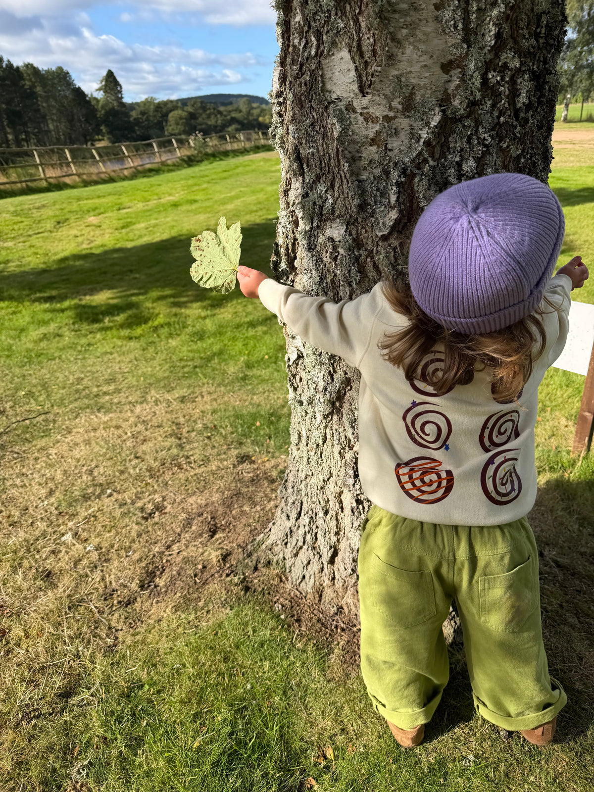 A toddler seen from behind stands in front of a tree outdoors, wearing a cream knit sweater with spiral motifs, olive green pants, and a lavender knit hat, while holding a light green leaf.