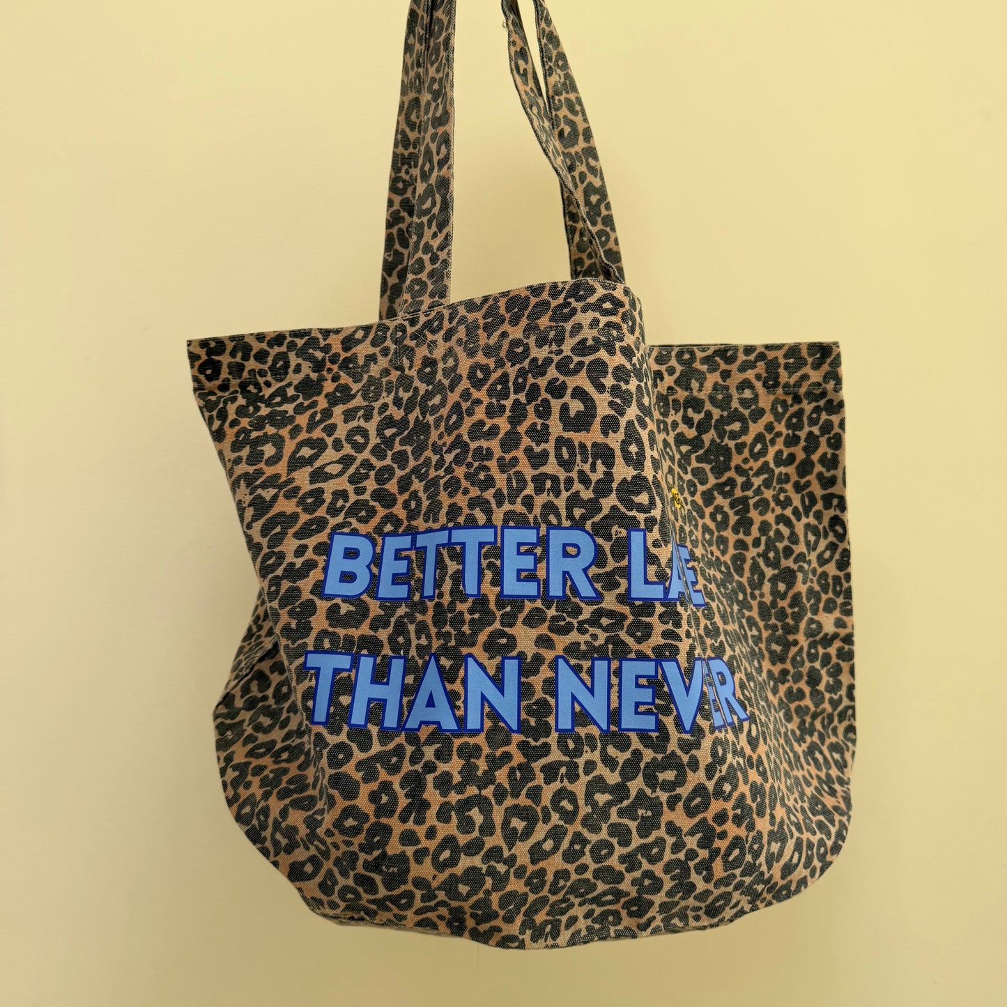 A leopard print tote bag with blue text reading 'BETTER LA THAN NEVA' being held by a hand against a plain beige wall.