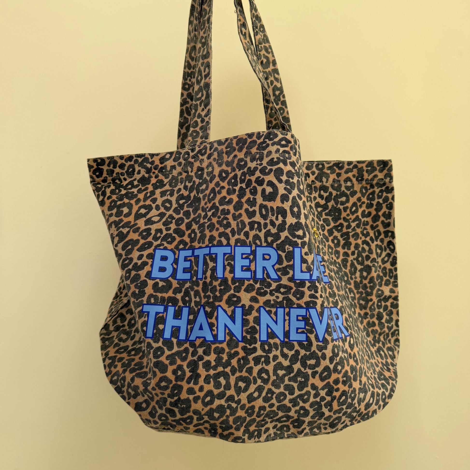 A leopard print tote bag with blue text reading 'BETTER LA THAN NEVA' being held by a hand against a plain beige wall.