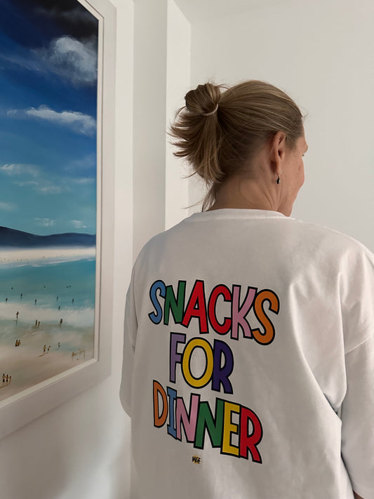 Person wearing a hoodie with 'Snacks for Dinner' text in colorful letters, standing against a white wall.
